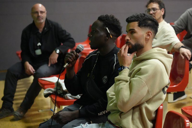 Un home jove negre parla amb un micròfon en una sala d'actes amb cadires vermelles de plàstic. Porta una dessuadora de xandall de la marca Jordan amb un logotip visible i té un bolígraf darrere l'orella. Al seu costat, un altre home jove amb una dessuadora beix s'està tocant la boca amb la mà, escoltant atentament amb una expressió pensativa. Al fons, diverses persones, inclòs un home calb amb ulleres, estan assegudes a les cadires vermelles.